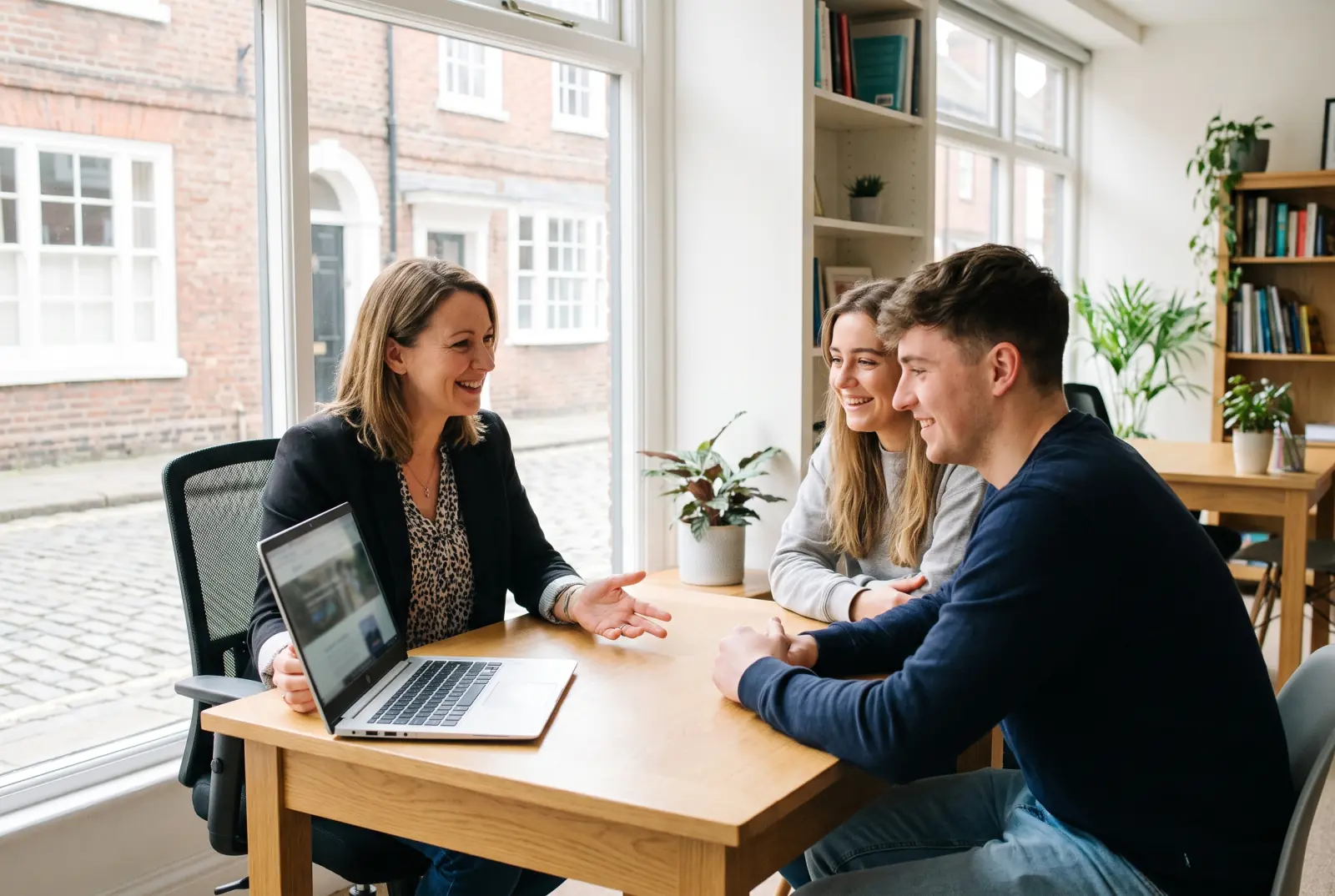 A mortgage adviser sitting across a table from a young couple, talking them through their options with a laptop open between them.