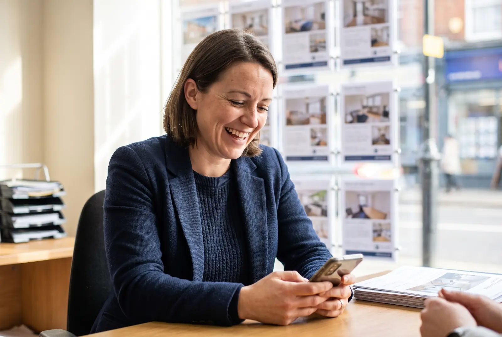 A warm, approachable British female letting agent in her early thirties at the desk of a bright UK high-street lettings branch, smiling as she looks at her phone mid-conversation, with a property window display behind her