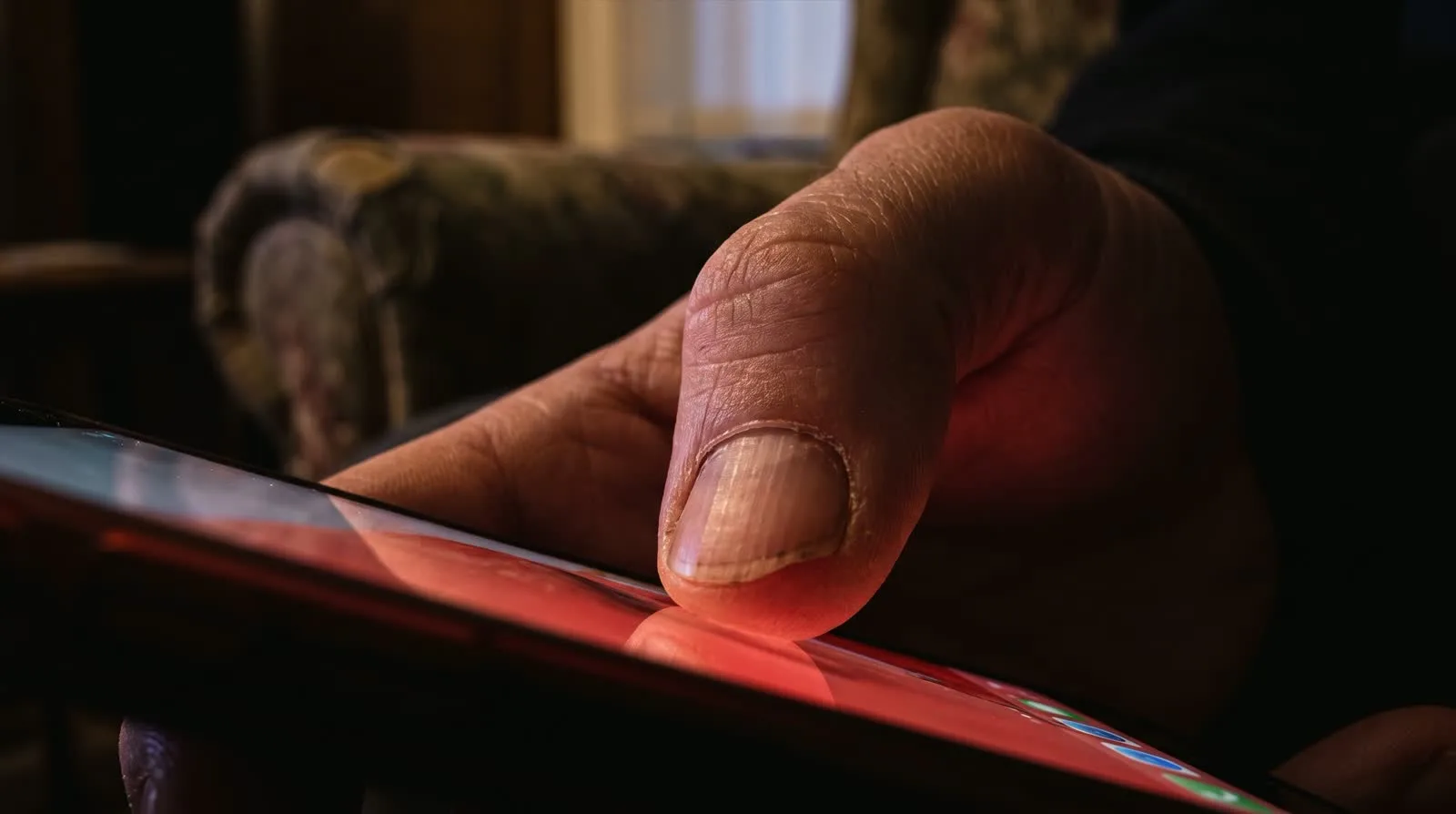A thumb lit red above the decline button of an incoming call on a phone screen, an armchair and a dark room behind